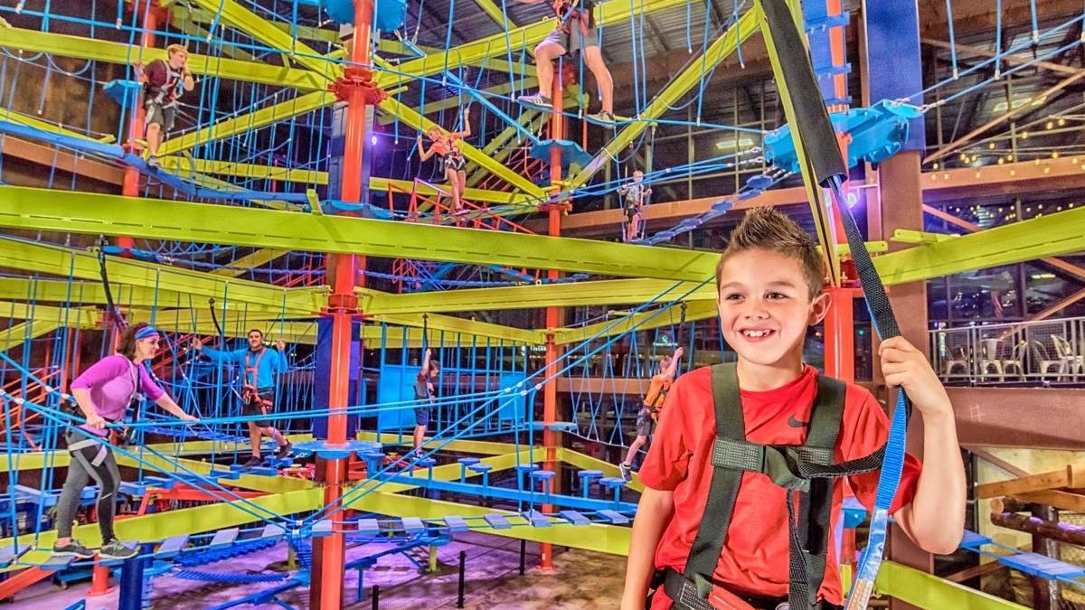 Small child in a red shirt with climbing gear on in the middle of a large climbing apparatus with other people climbing around him